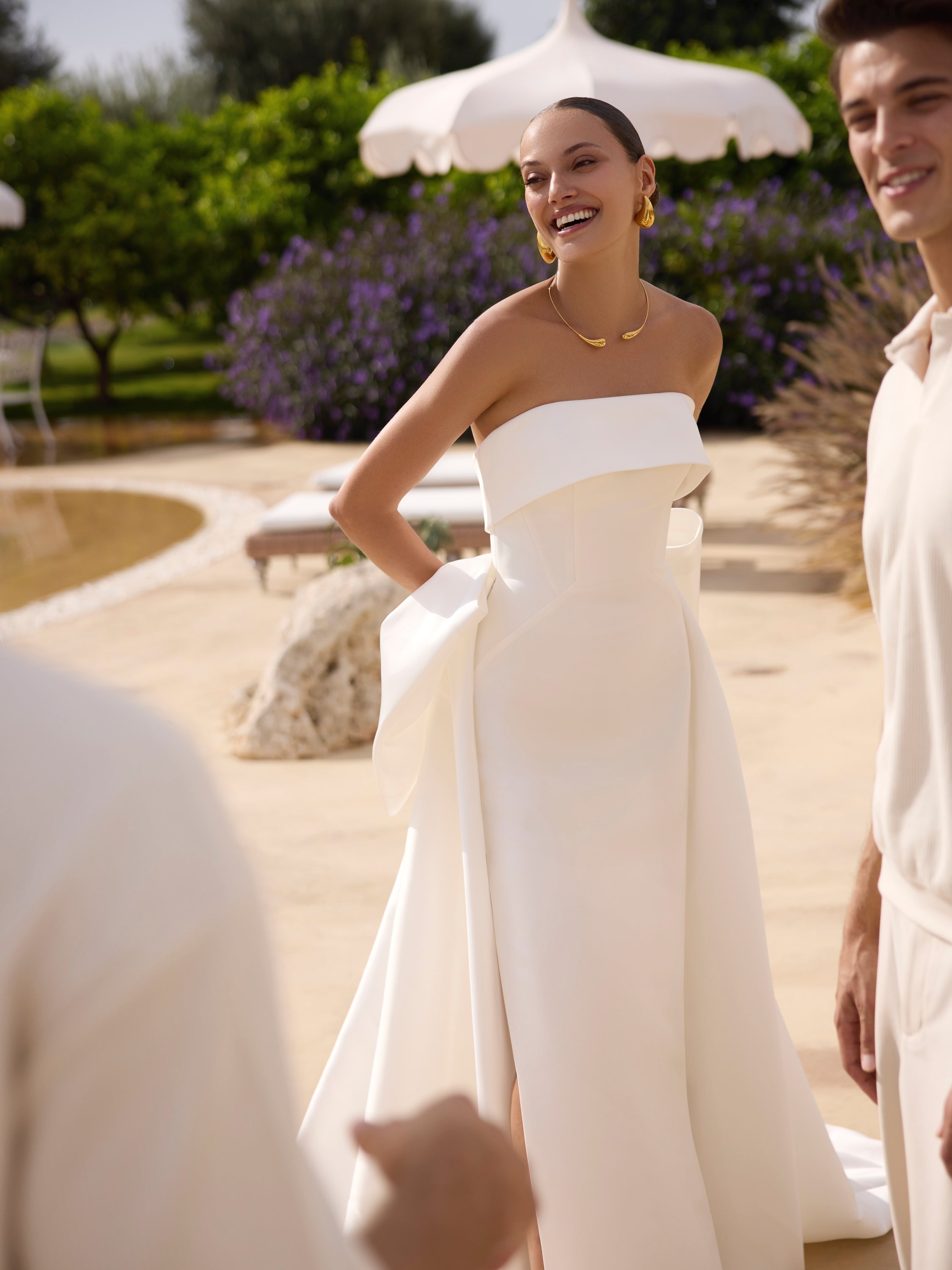 Woman in Genesis wedding dress by Modeca standing outdoors with greenery and a white umbrella in the background.