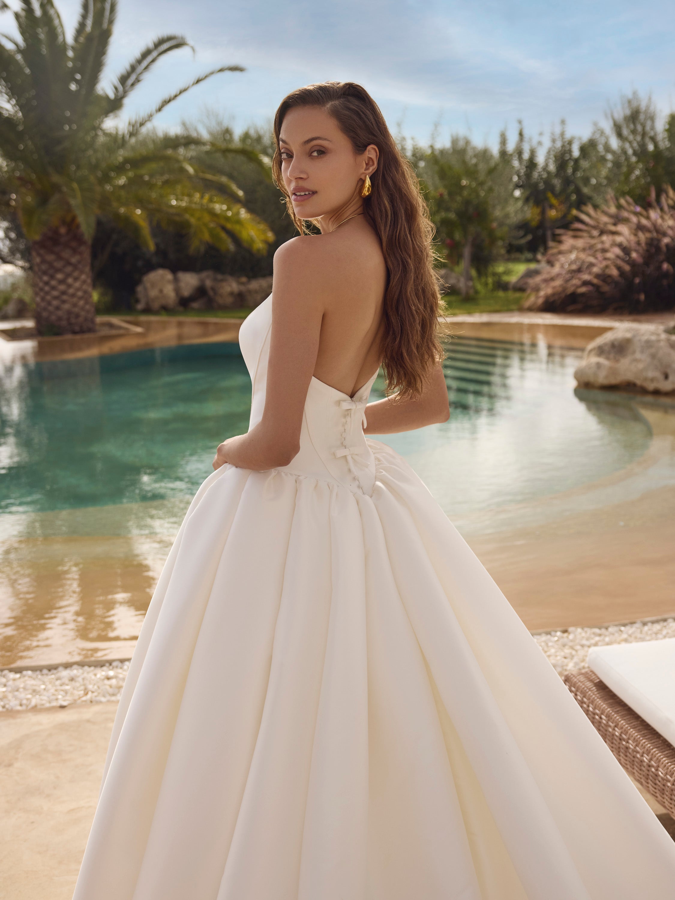 Woman in Ghada wedding dress by Modeca standing by a pool with palm trees in the background