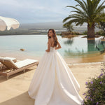 Woman in Ghada wedding dress by Modeca standing by a pool with palm trees and a clear sky.