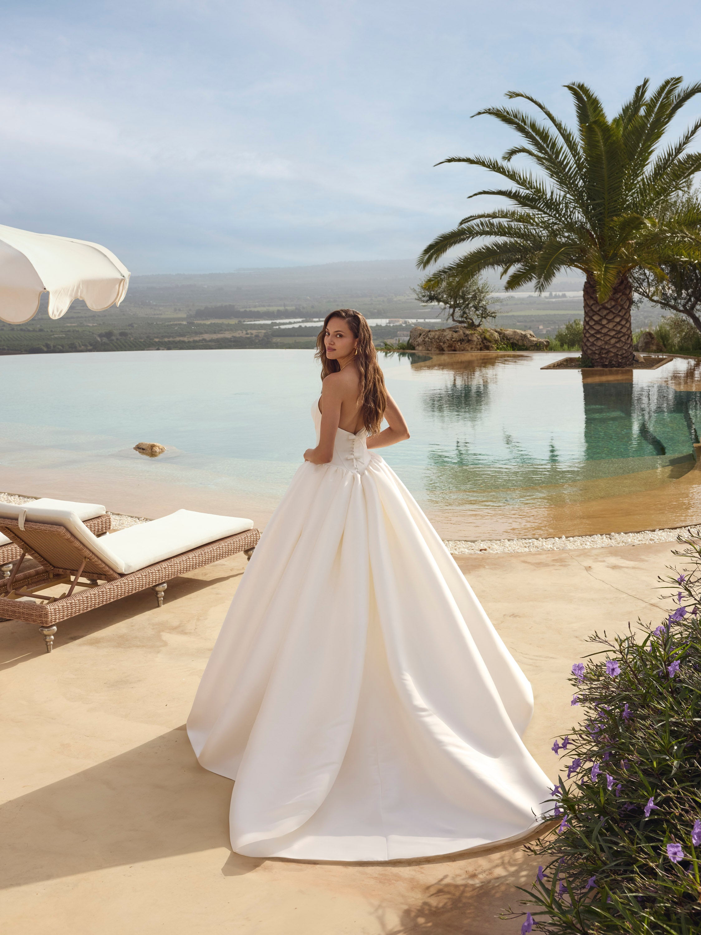 Woman in Ghada wedding dress by Modeca standing by a pool with palm trees and a clear sky.