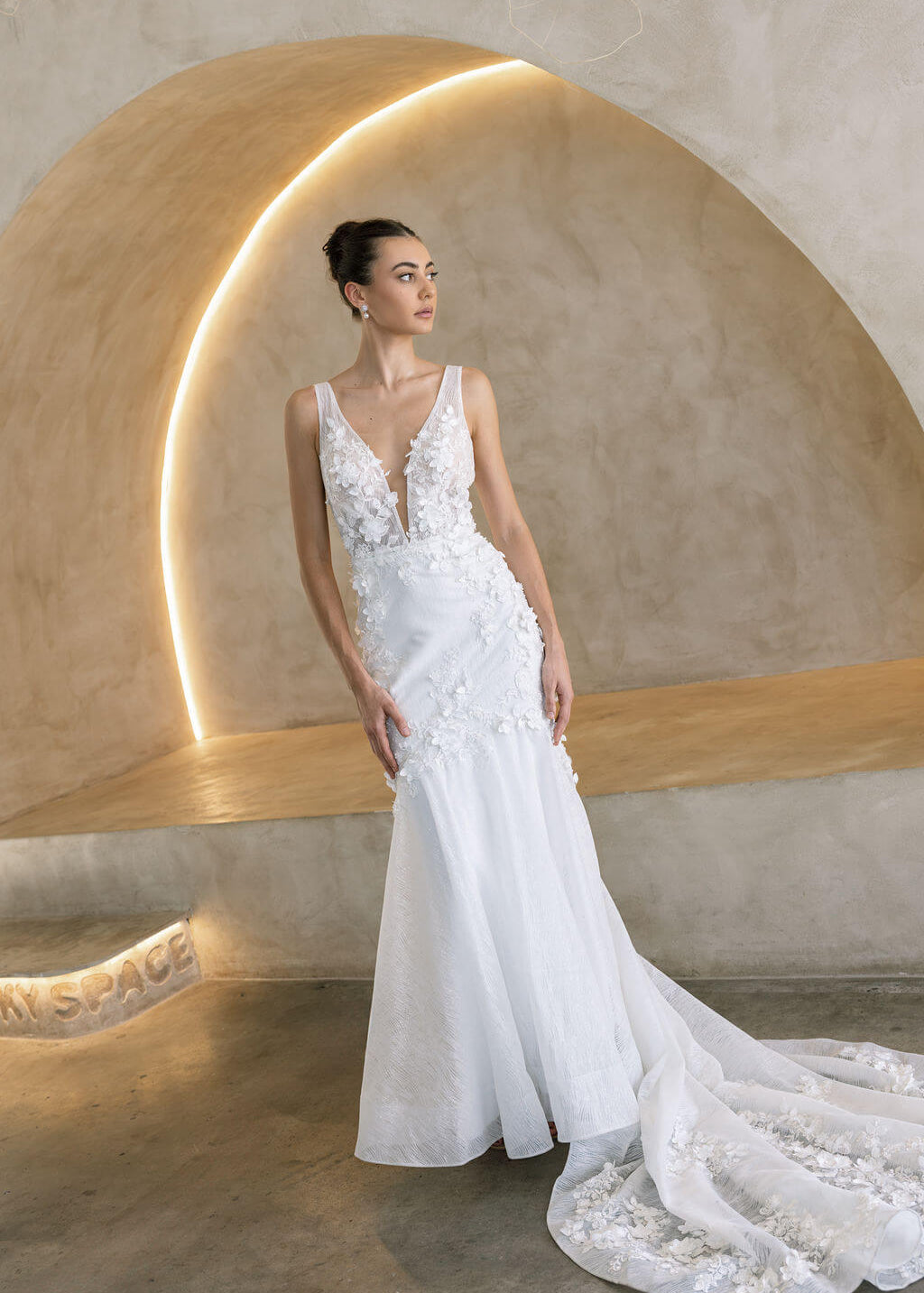 Woman in a Floral Fit and Flare Bridal Gown standing against a textured wall with architectural elements.