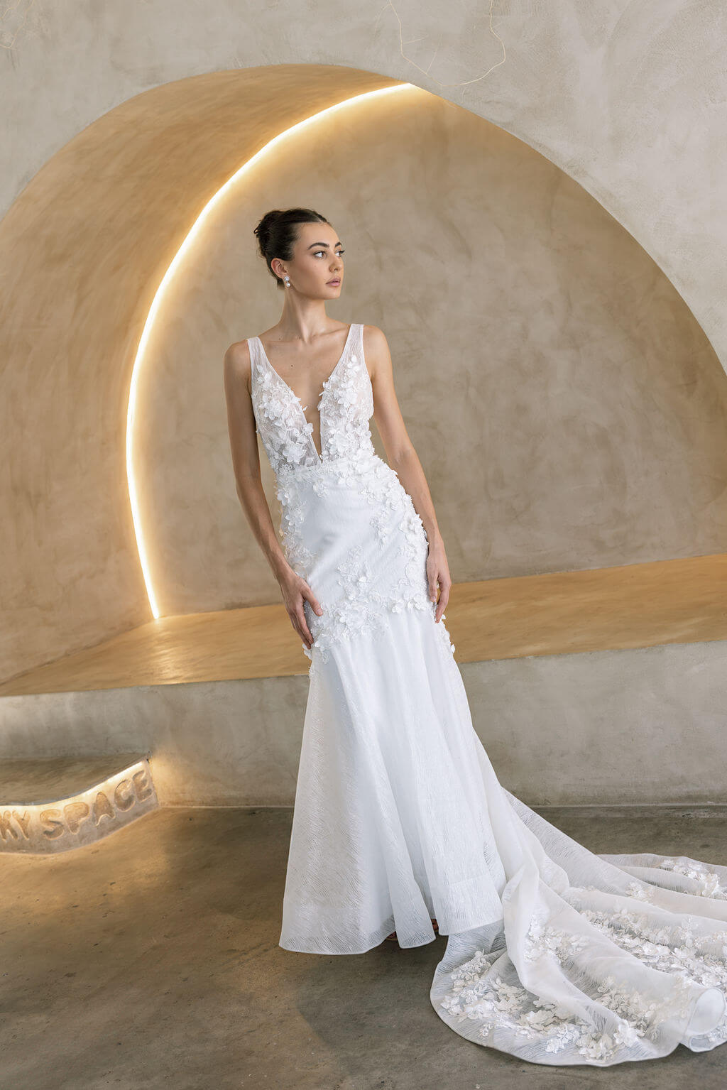 Woman in a Floral Fit and Flare Bridal Gown standing against a textured wall with architectural elements.