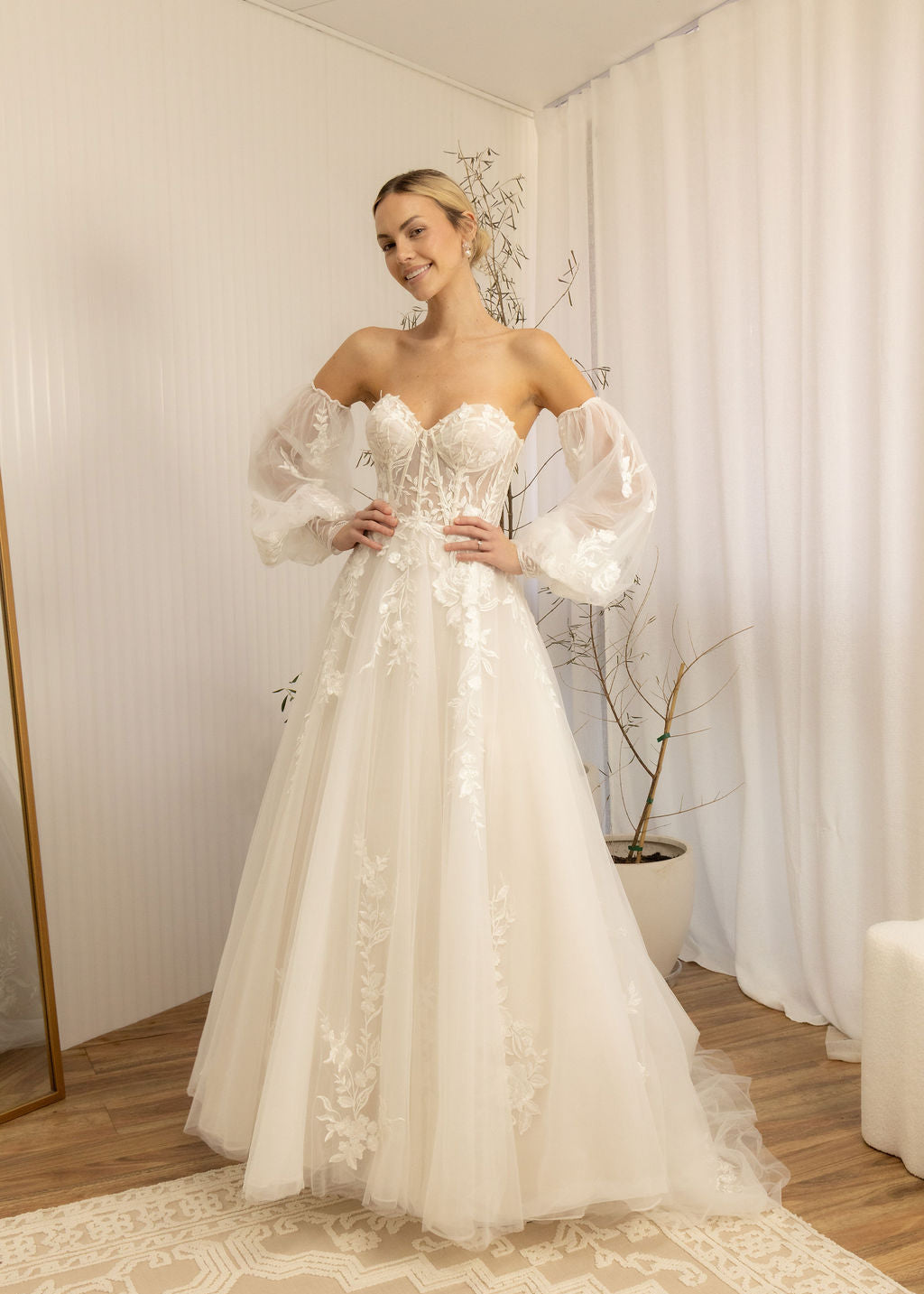 Woman wearing a white Romantic Lace Wedding Dress in a room with curtains and a plant.