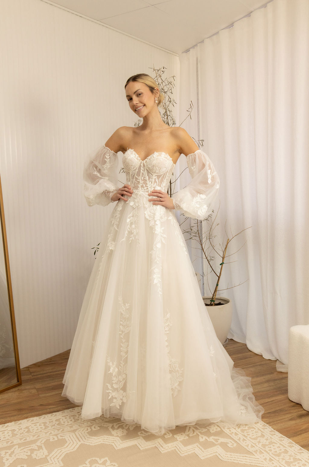 Woman wearing a white Romantic Lace Wedding Dress in a room with curtains and a plant.