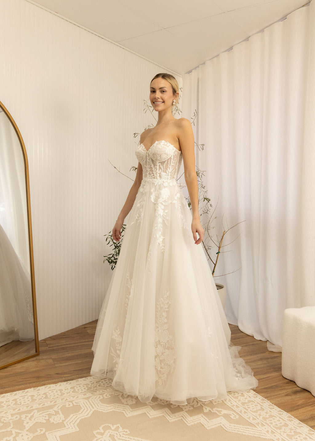 Woman in a white Romantic Lace Wedding Dress standing in a room with white walls and a white curtain.