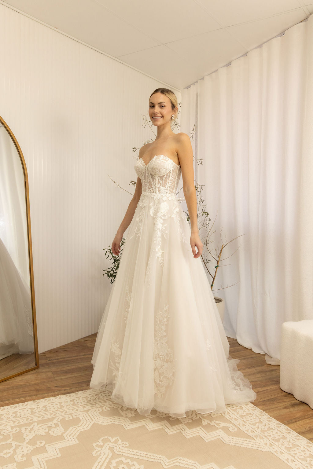 Woman in a white Romantic Lace Wedding Dress standing in a room with white walls and a white curtain.