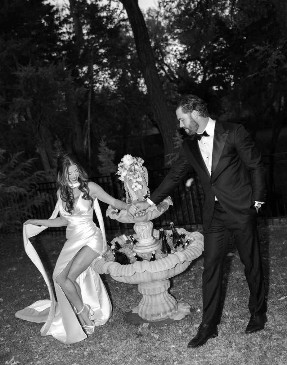 Bride wearing the Heirloom modern A-line wedding dress with leg split and veil, standing with groom beside a garden fountain.