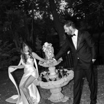 Bride wearing the Heirloom modern A-line wedding dress with leg split and veil, standing with groom beside a garden fountain.