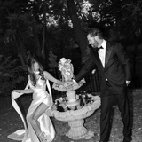 Bride wearing the Heirloom modern A-line wedding dress with leg split and veil, standing with groom beside a garden fountain.