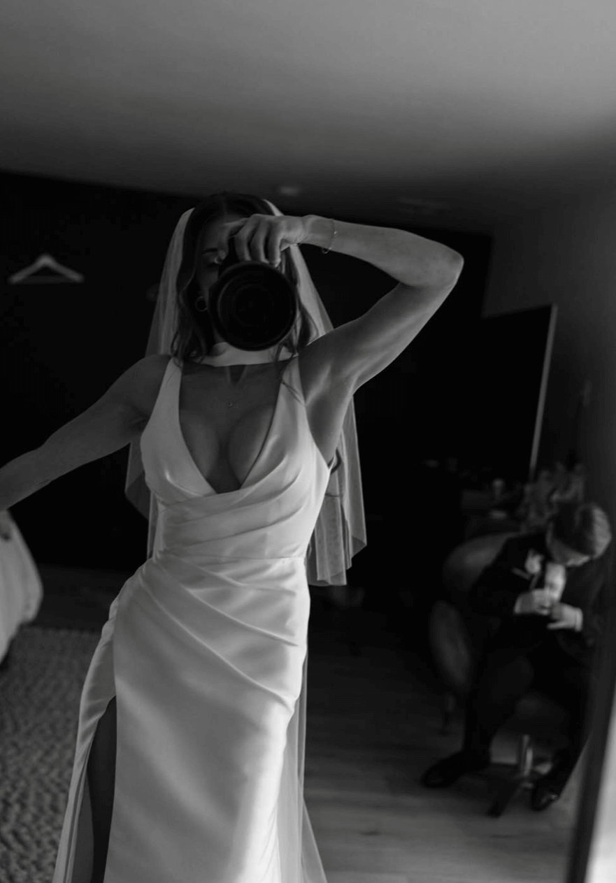 Bride wearing the Heirloom modern A-line wedding dress with veil and leg split, taking a mirror photo while holding a camera.