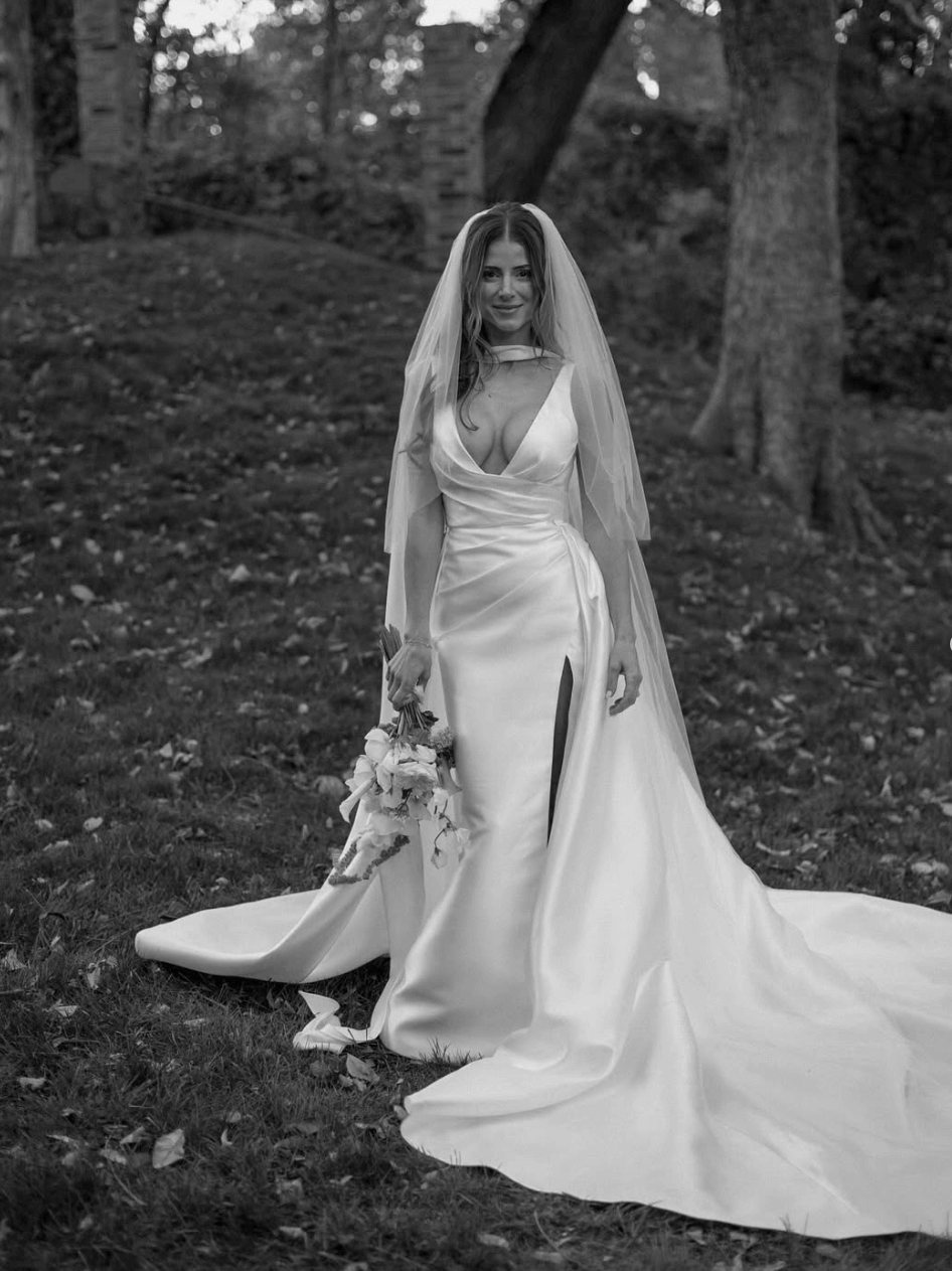 Bride wearing the Heirloom modern A-line wedding dress with veil and leg split, holding a bouquet in an outdoor garden setting.