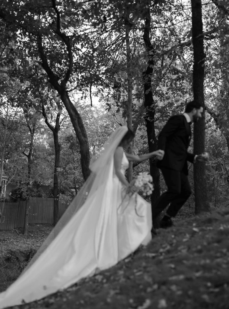 Bride wearing the Heirloom modern A-line wedding dress with veil and long train, walking with groom through a wooded outdoor setting.