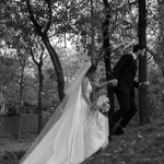 Bride wearing the Heirloom modern A-line wedding dress with veil and long train, walking with groom through a wooded outdoor setting.