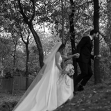 Bride wearing the Heirloom modern A-line wedding dress with veil and long train, walking with groom through a wooded outdoor setting.