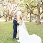Couple in wedding attire embracing in a park with trees and grass.