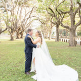 Couple in wedding attire embracing in a park with trees and grass.