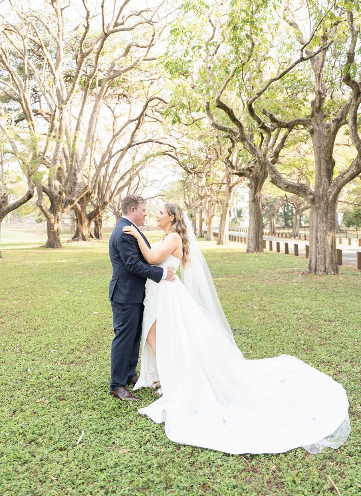 Couple in wedding attire embracing in a park with trees and grass.