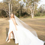 Woman in a Fit and Flare Wedding Dress with Overskirt with a long veil standing in a grassy field with trees.
