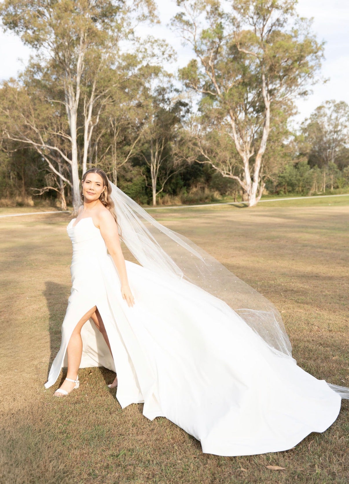 Woman in a Fit and Flare Wedding Dress with Overskirt with a long veil standing in a grassy field with trees.