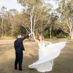 Man lifting a woman in a Fit and Flare Wedding Dress with Overskirt in an outdoor setting with trees.