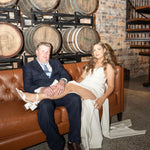 Man and woman sitting on a brown leather couch in front of wine barrels with a brick wall background.