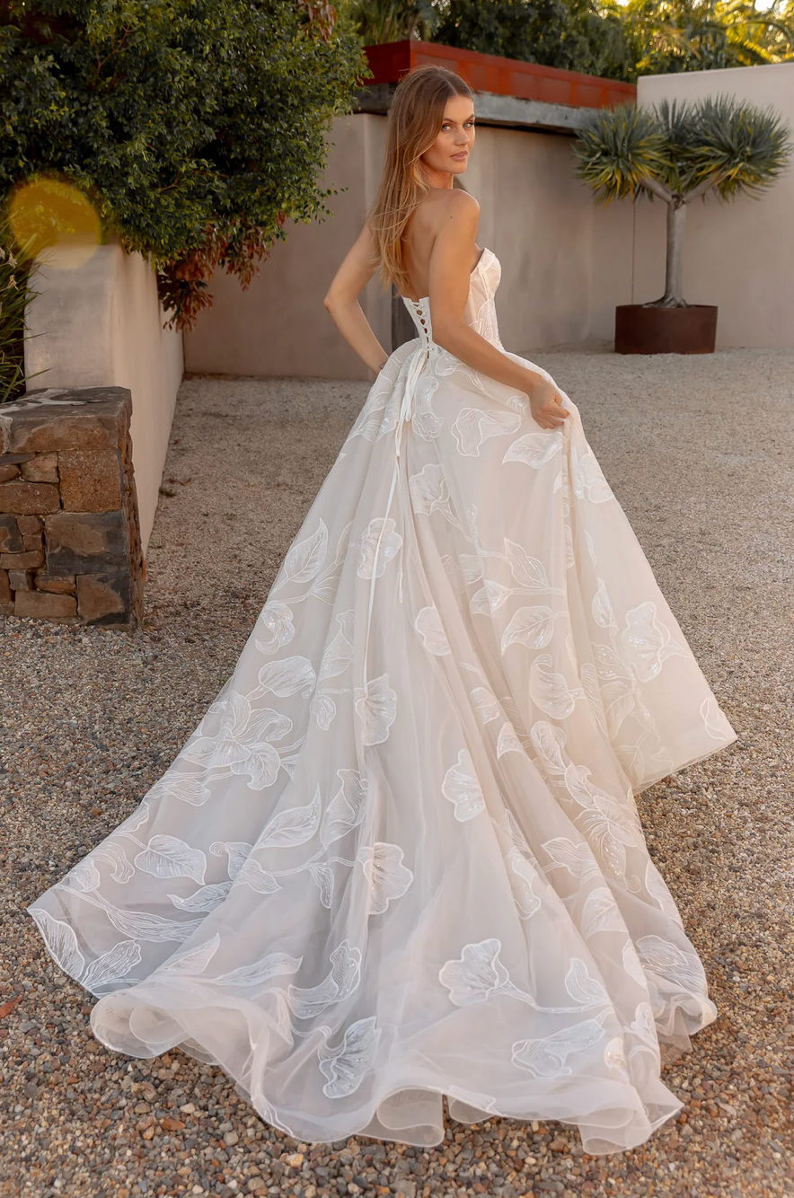 Woman in an after five lace appliqué A-line wedding dress standing outdoors with plants and stone wall in the background