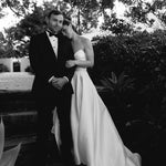 Black and white photo of a couple in formal attire standing outdoors.Bride wearing Marigold Mikado A-line wedding dress with strapless sweetheart bodice standing with groom in a garden setting.