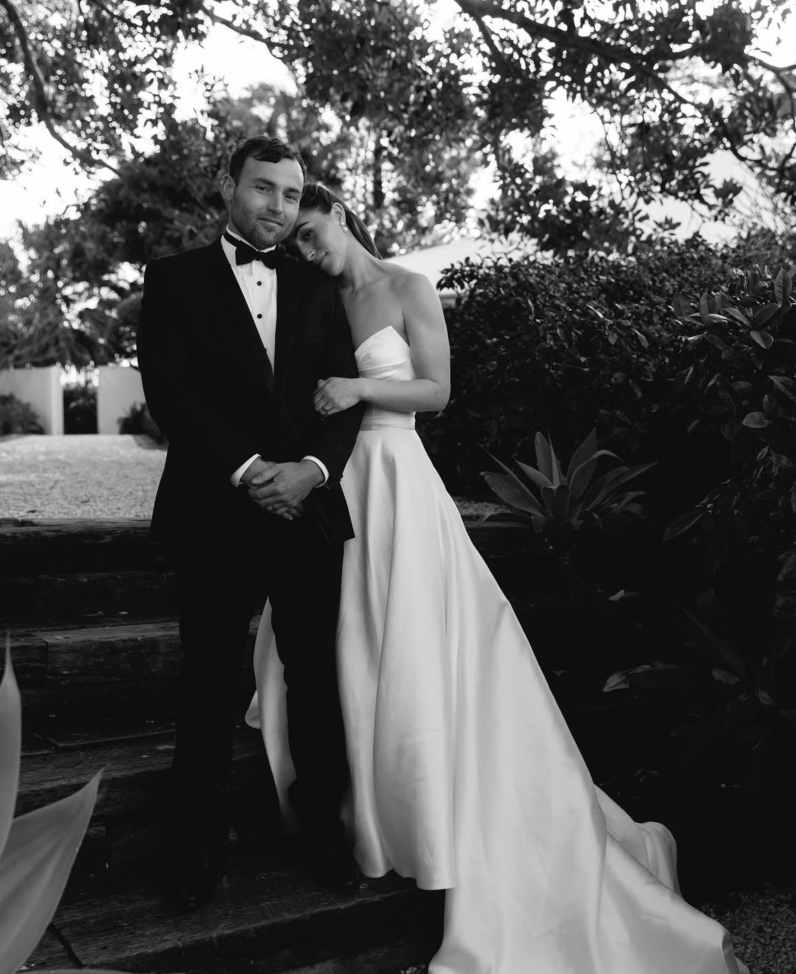 Black and white photo of a couple in formal attire standing outdoors.Bride wearing Marigold Mikado A-line wedding dress with strapless sweetheart bodice standing with groom in a garden setting.
