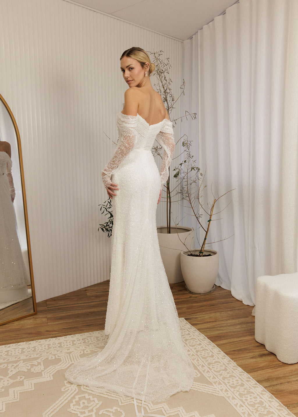 Woman in a Off Shoulder Fitted Wedding Dress standing in a room with a mirror and plants.