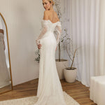 Woman in a Off Shoulder Fitted Wedding Dress standing in a room with a mirror and plants.