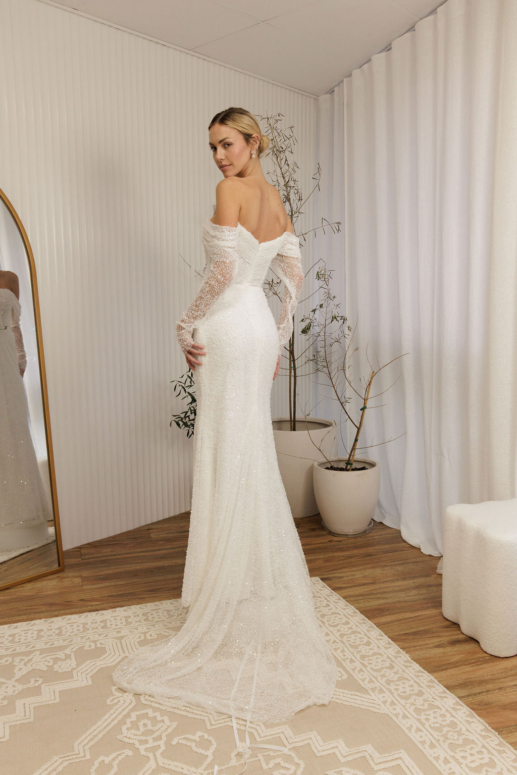 Woman in a Off Shoulder Fitted Wedding Dress standing in a room with a mirror and plants.
