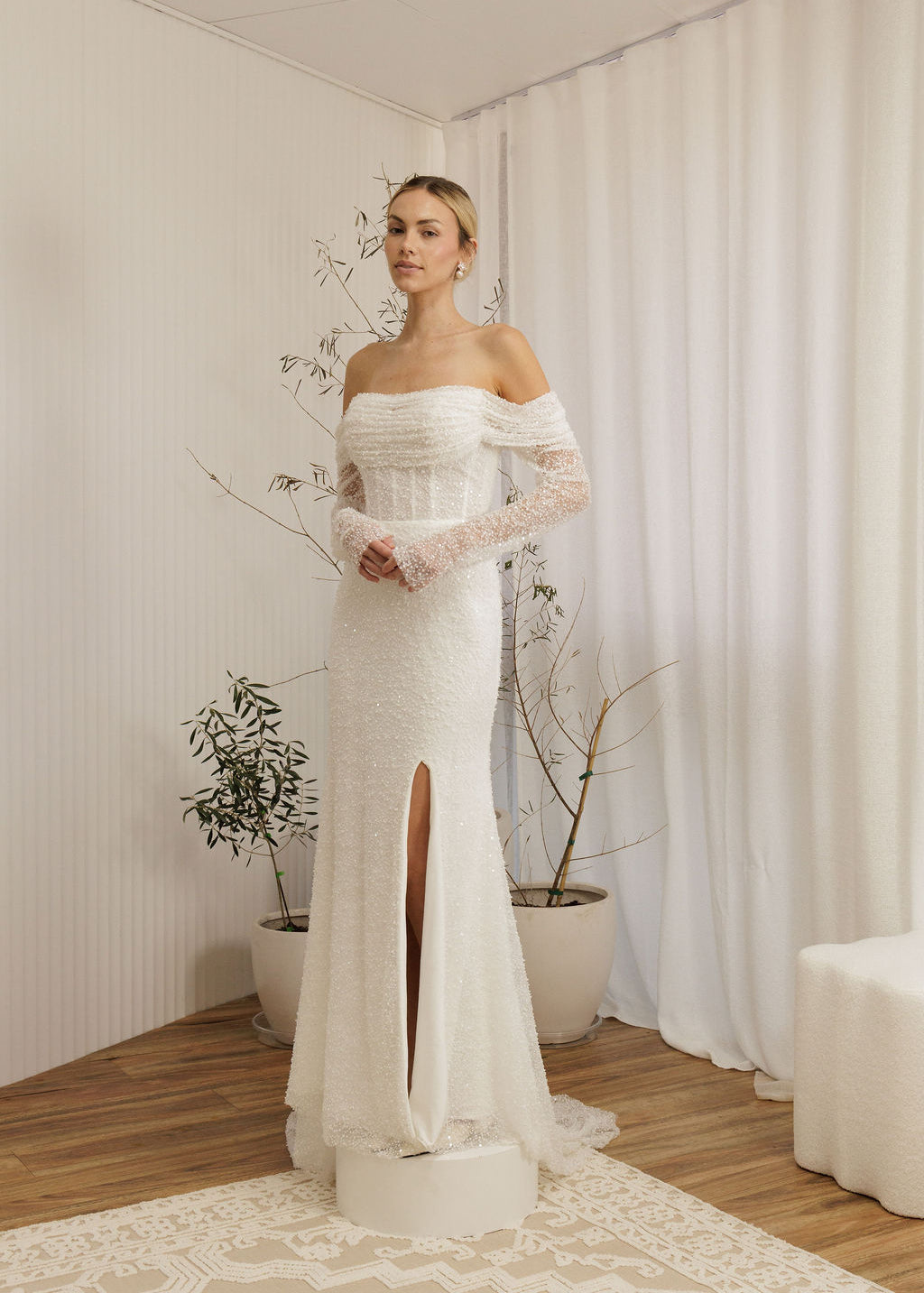 Woman wearing a Off Shoulder Fitted Wedding Dress with a high slit in a room with white curtains and plants.