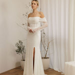 Woman wearing a Off Shoulder Fitted Wedding Dress with a high slit in a room with white curtains and plants.