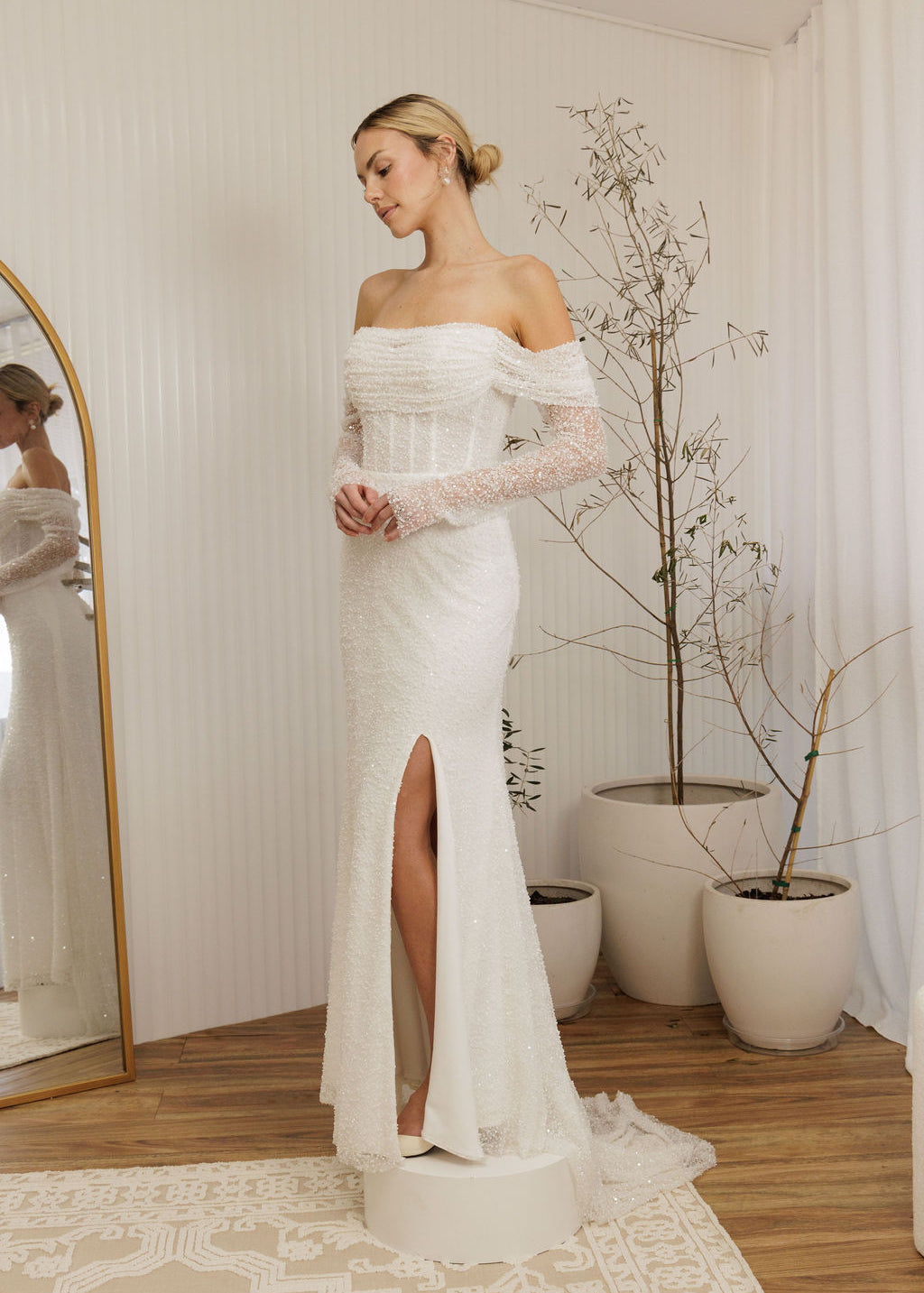 Woman in a Off Shoulder Fitted Wedding Dress standing in a room with a mirror and plants.