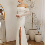 Woman in a Off Shoulder Fitted Wedding Dress standing in a room with a mirror and plants.