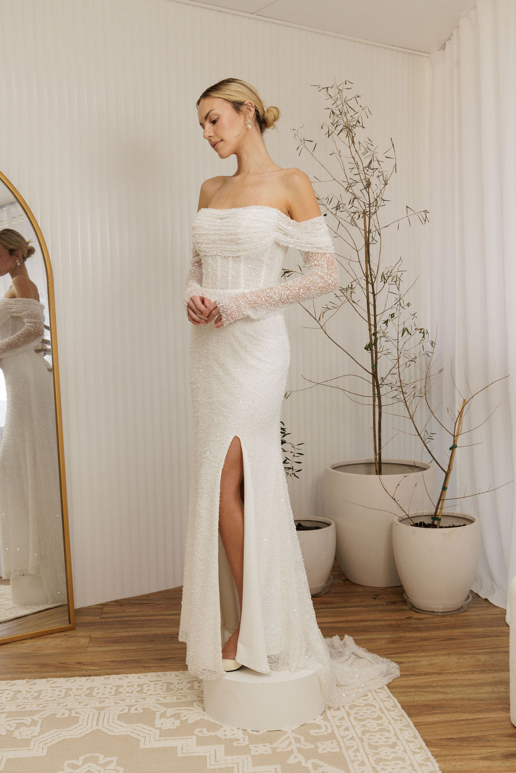 Woman in a Off Shoulder Fitted Wedding Dress standing in a room with a mirror and plants.