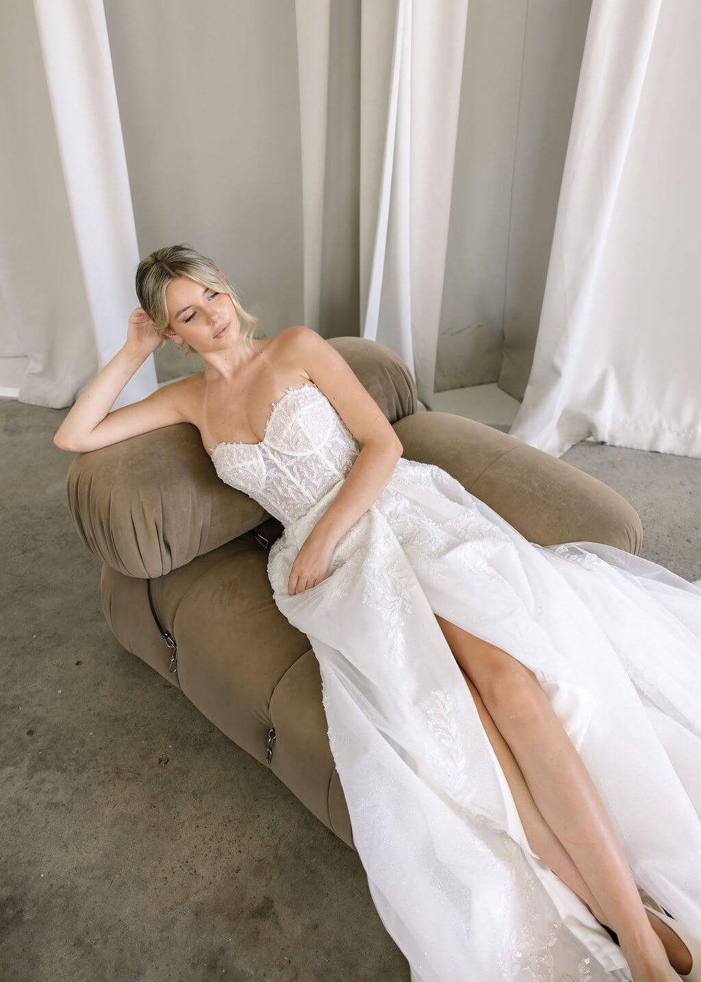 Woman in a Lace A-Line Wedding Dress sitting on a brown couch with white curtains in the background.