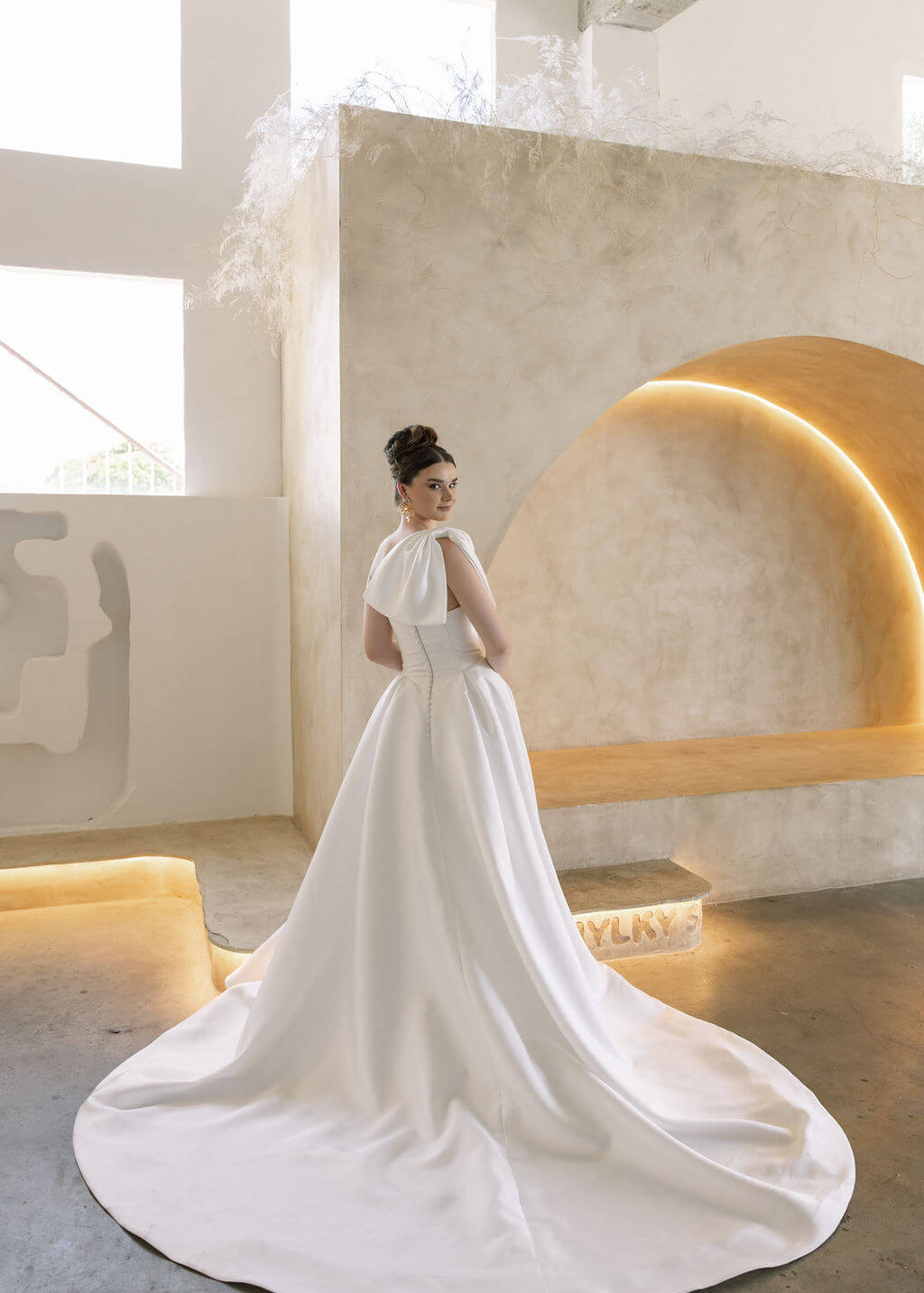 Woman in a white modern mikado wedding dress standing in a modern indoor setting with a large mirror.