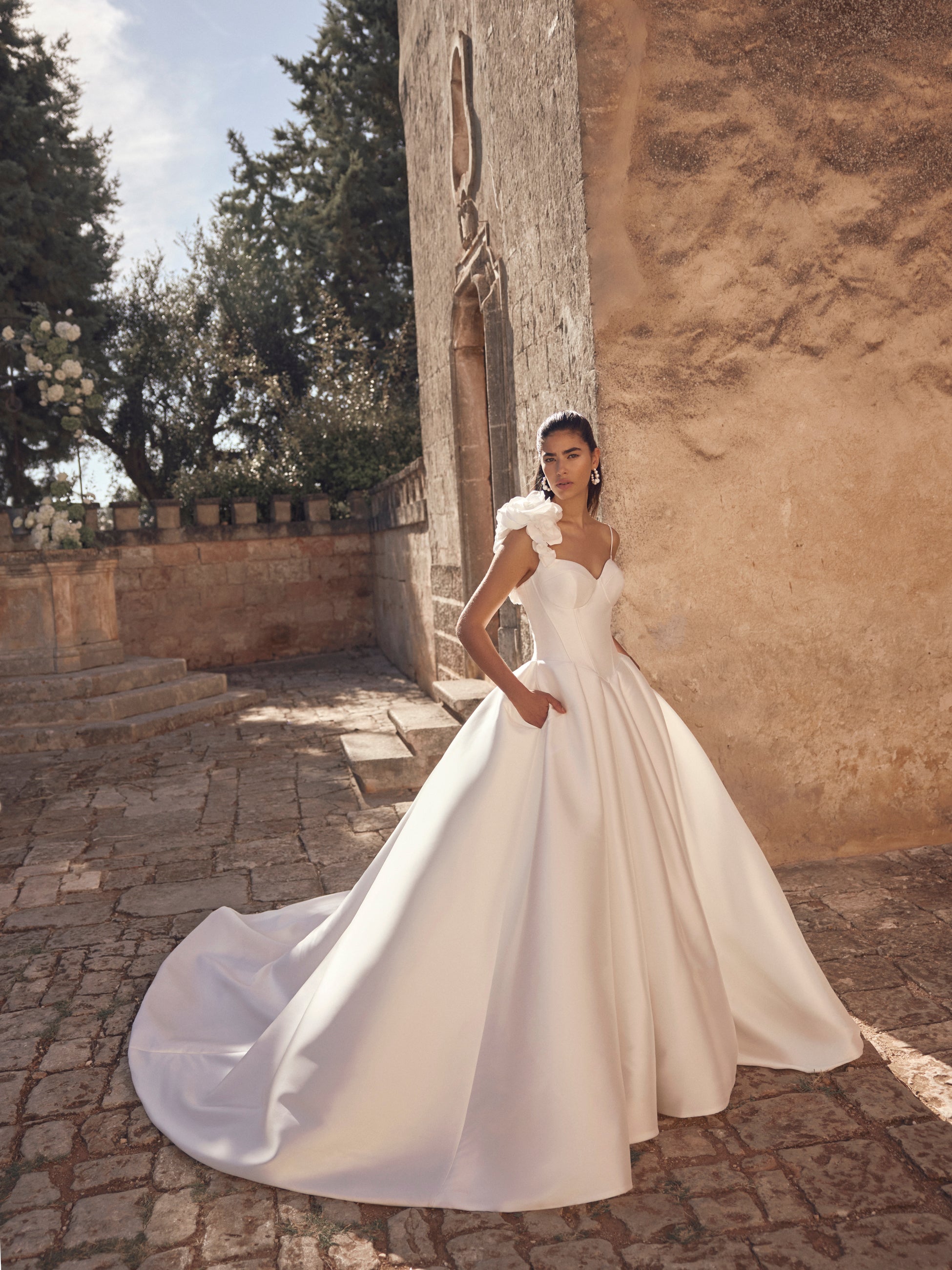 A woman wearing a voluminous white wedding gown stands outdoors beside a rustic stone wall and cobblestone path. The dress has a fitted bodice, dramatic off-shoulder ruffle details, and a full flowing skirt with a long train.