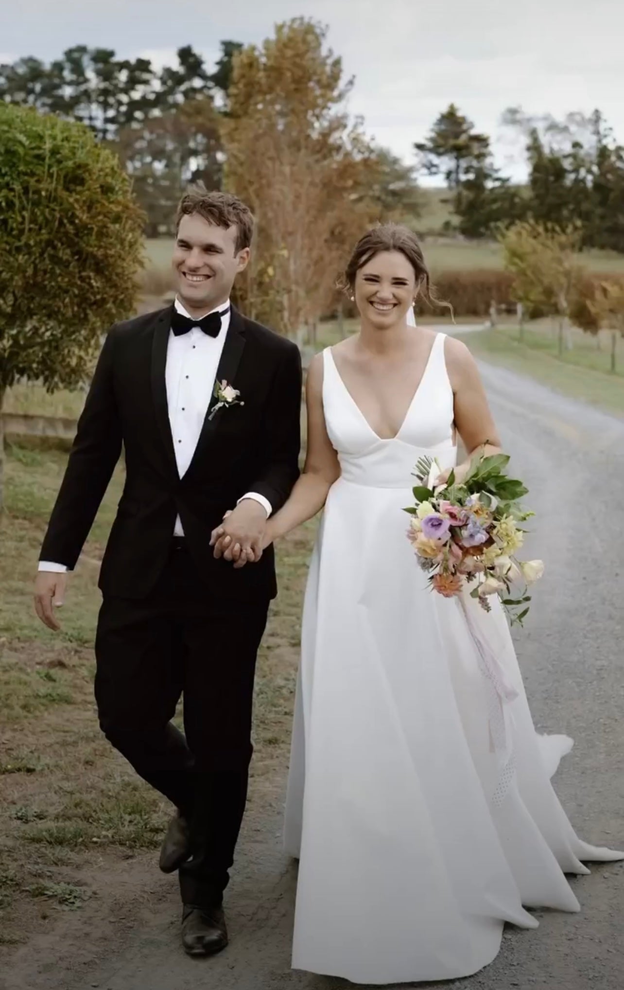 Bride wearing the Phoenix plunge V-neck wedding dress walking with groom along a country road, holding a bouquet.