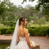 Bride in a white wedding dress with a long veil standing by a pond with greenery in the background.