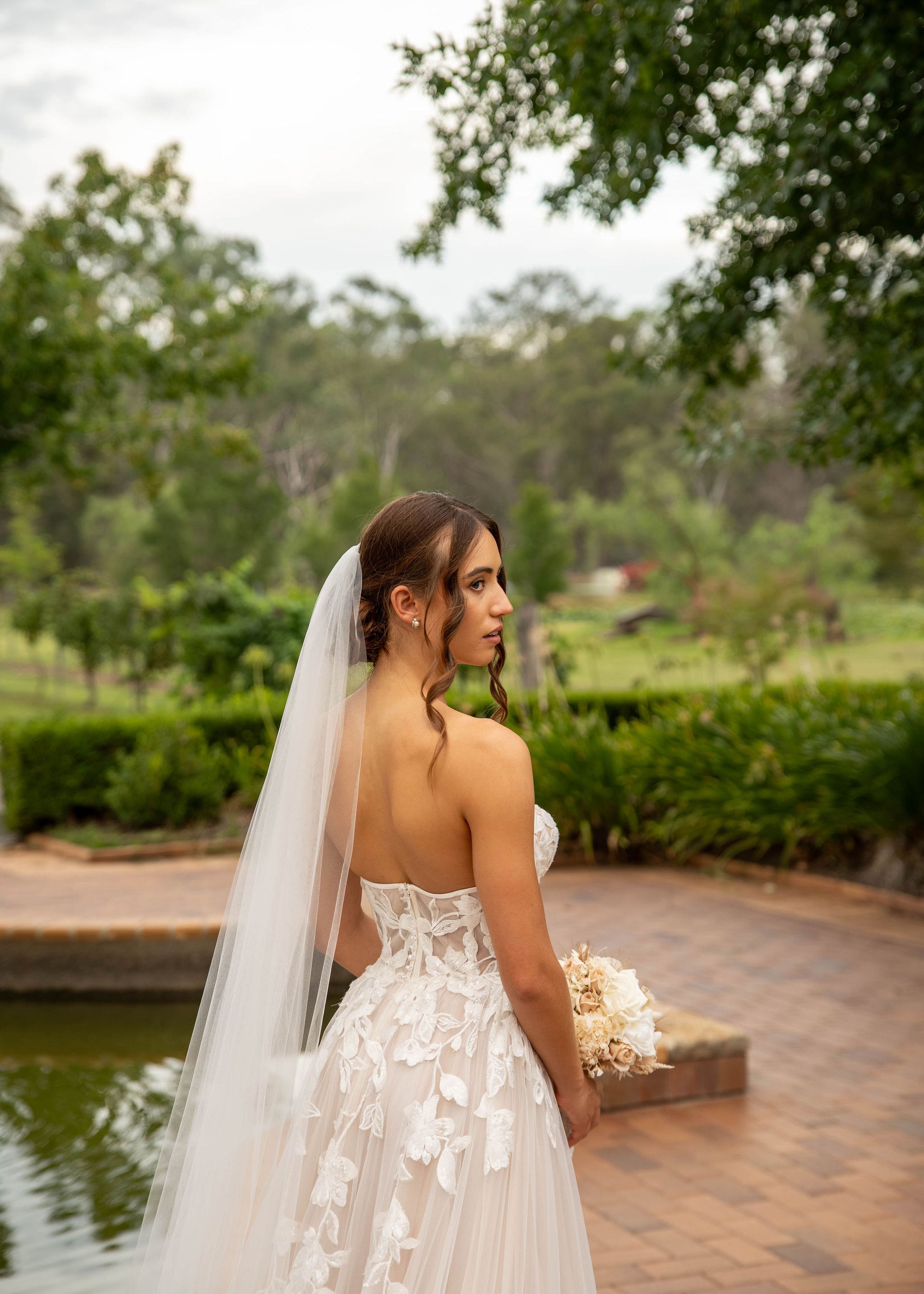 Bride in a white wedding dress with a long veil standing by a pond with greenery in the background.