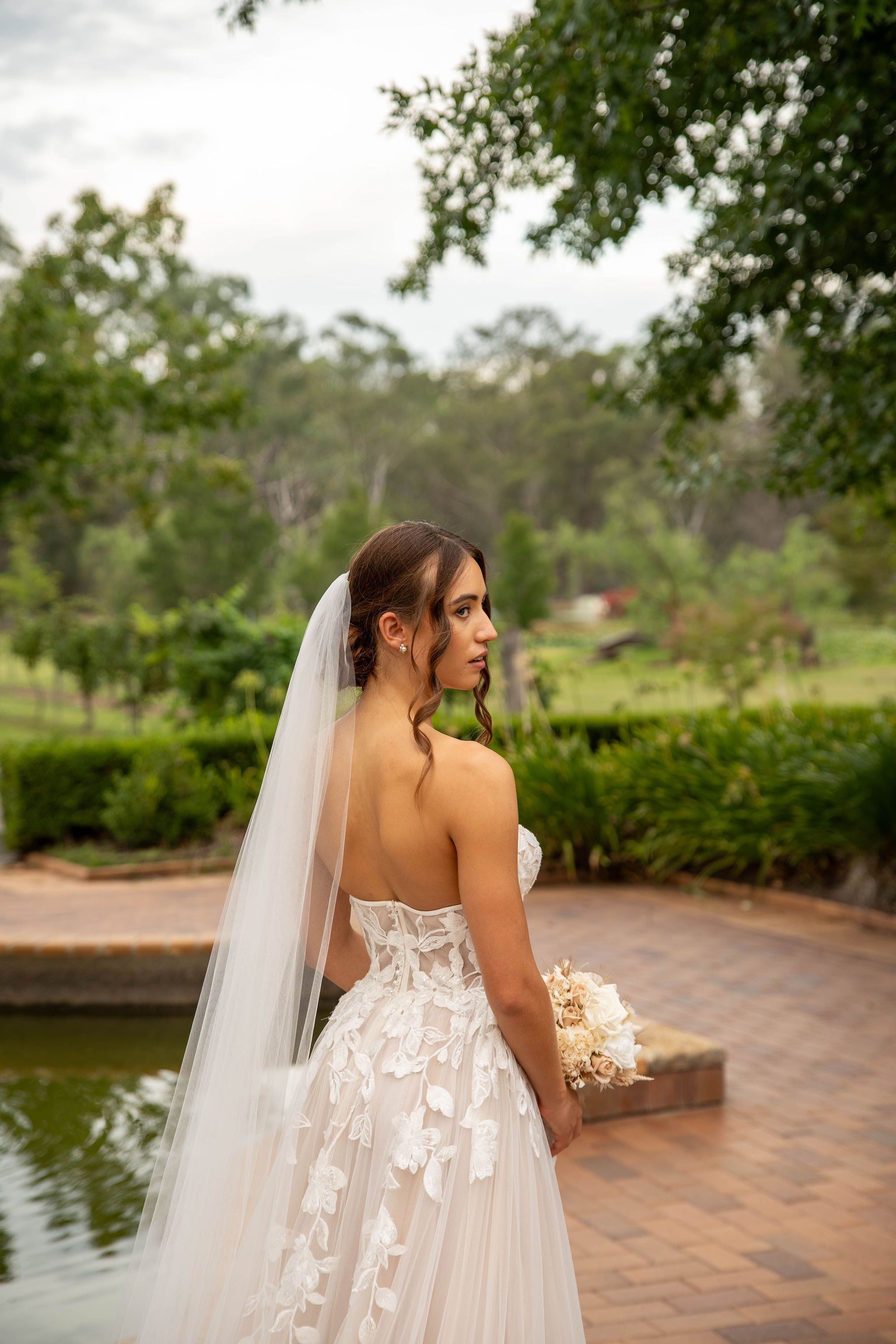Bride in a white wedding dress with a long veil standing by a pond with greenery in the background.