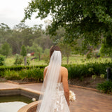 Bride in a strapless gown with a long veil standing by a pool with greenery in the background