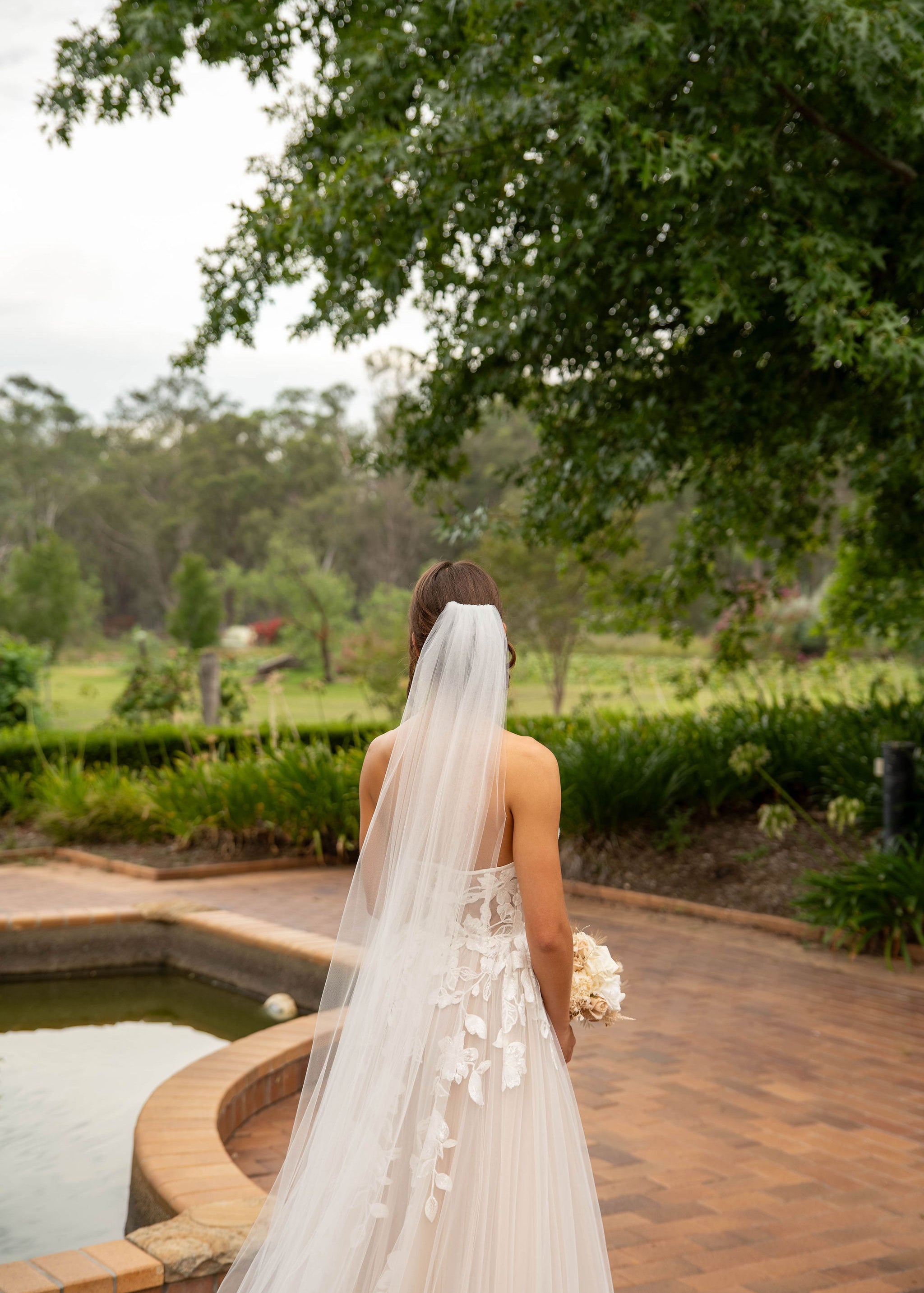 Bride in a strapless gown with a long veil standing by a pool with greenery in the background
