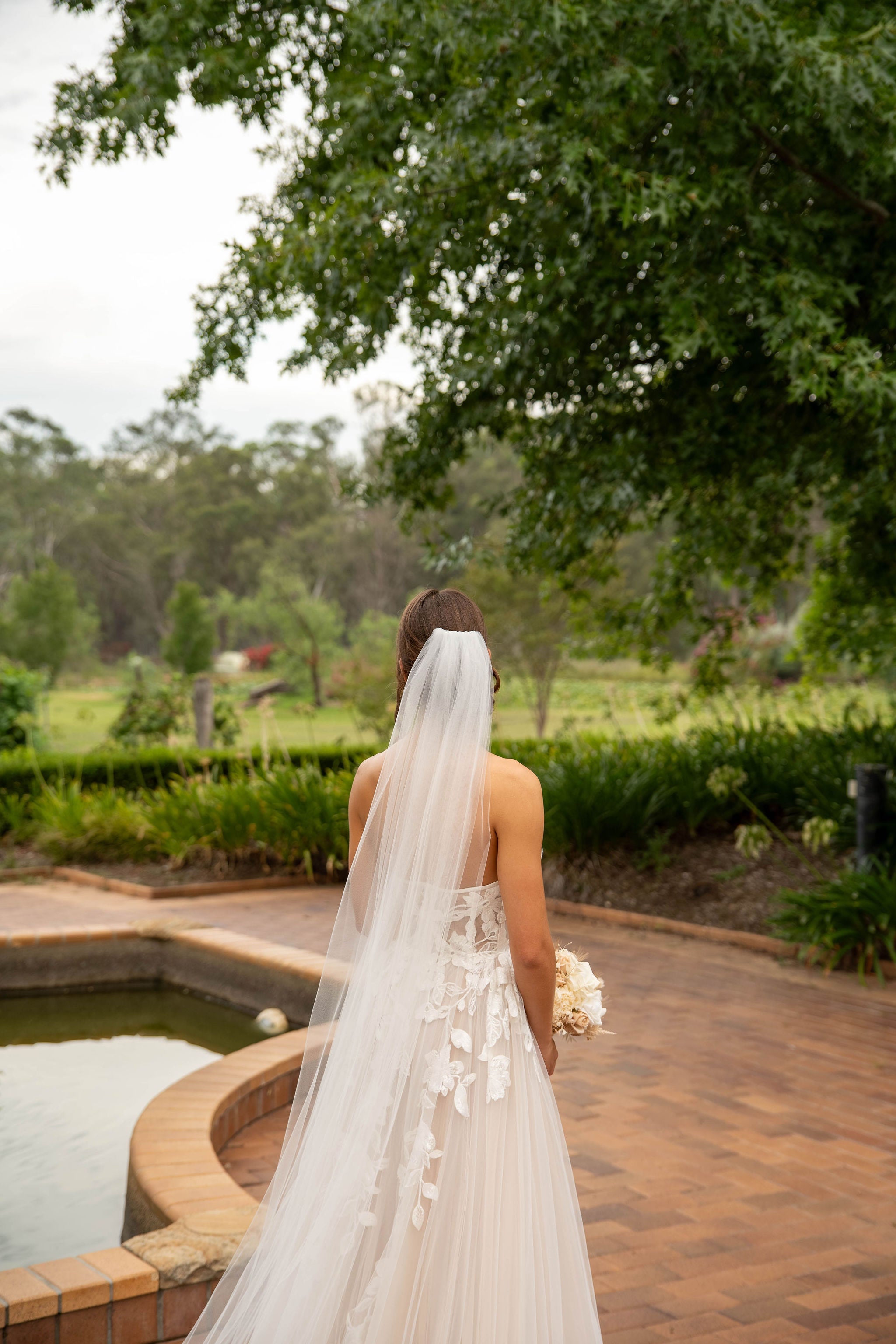 Bride in a strapless gown with a long veil standing by a pool with greenery in the background