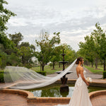 Woman in a wedding dress with a long veil standing by a fountain in a park.