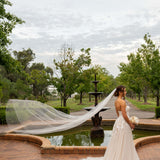 Woman in a wedding dress with a long veil standing by a fountain in a park.