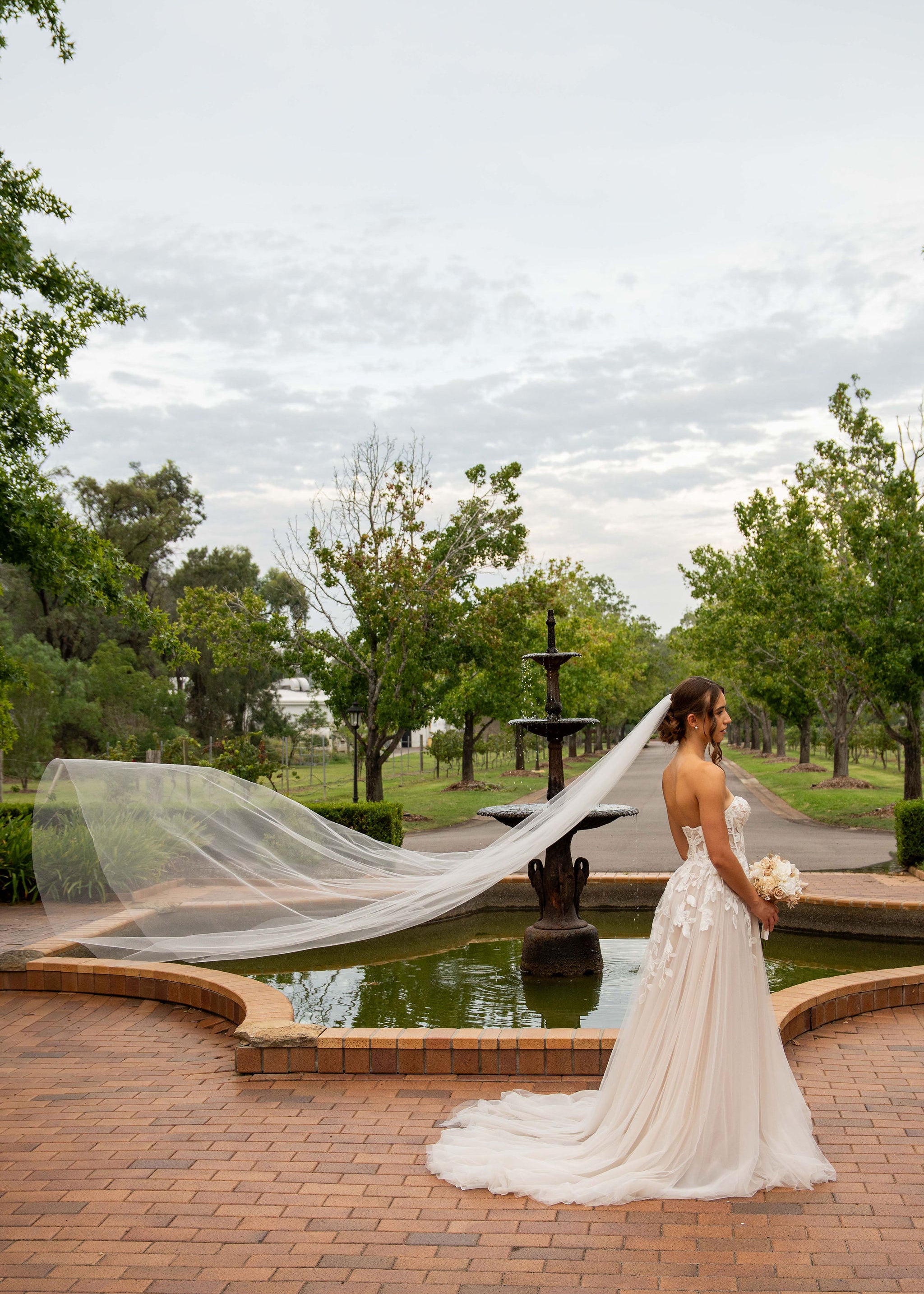Woman in a wedding dress with a long veil standing by a fountain in a park.