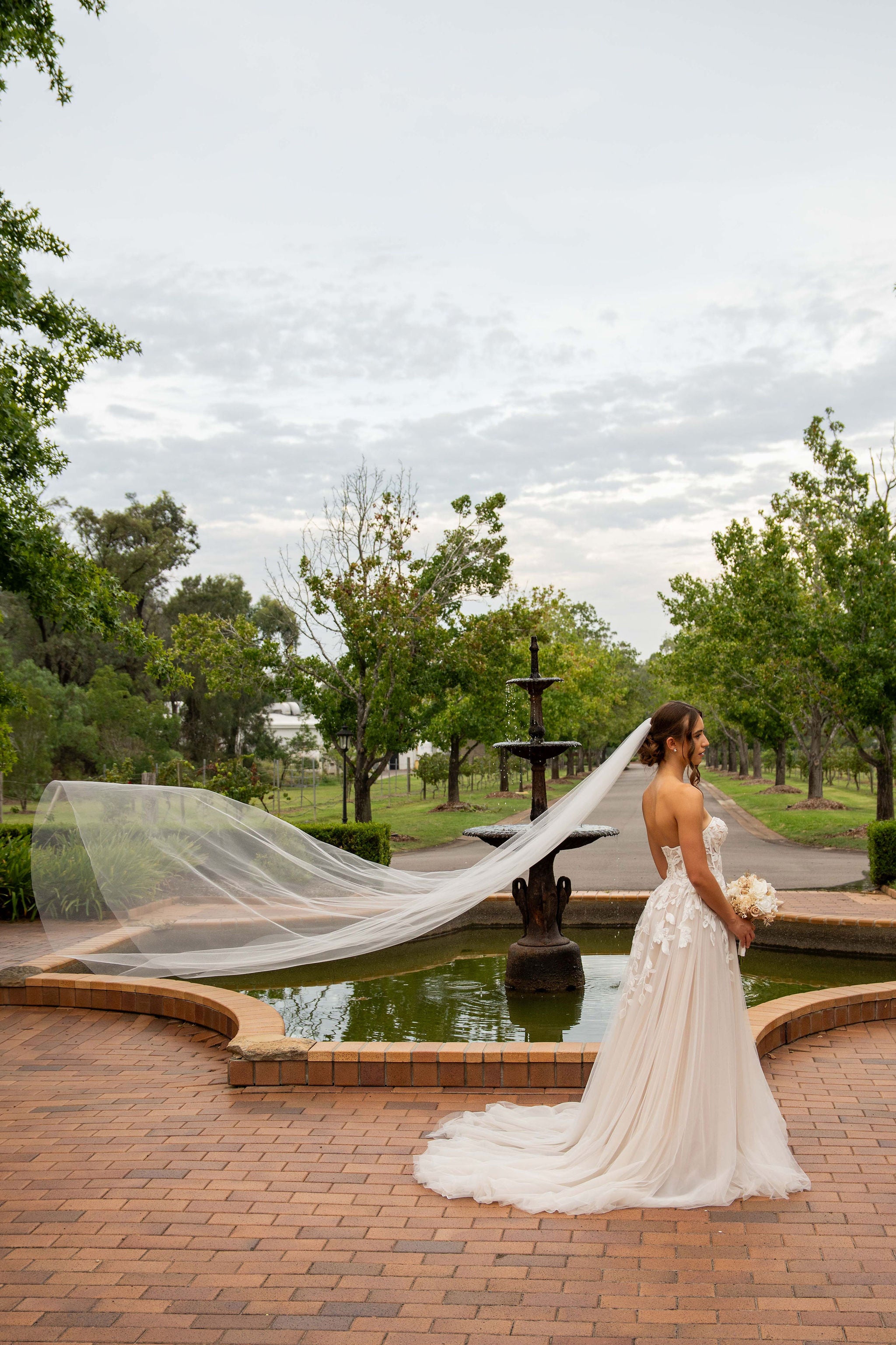 Woman in a wedding dress with a long veil standing by a fountain in a park.